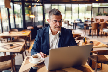 Middle aged Caucasian businessman with serious facial expression using laptop while sitting in cafe. On the desk coffee, in ears earphones.