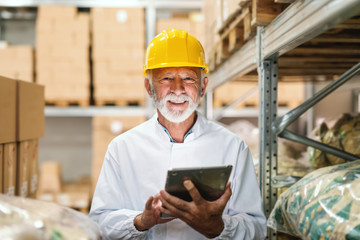 Smiling senior worker in uniform and with yellow helmet on head holding tablet and looking at camera while standing in storage.