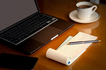 Mock up image of laptop computer,  blank checkbook, pen, phone and cup of coffee on wooden table background. Table setting,paycheck and business financial concept.