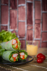 Salad rolls on black plate placed on a wood sieve there is tomatos placed beside and salad dressing placed backside.
