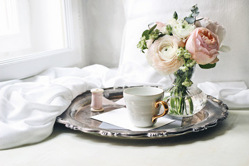 Spring, summer still life scene. Cup of coffee, blank greeting card, envelope on old silver tray at windowsill. Vintage feminine styled photo. Floral composition, pink English roses and Ranunculus.