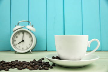 White coffee cup and coffee bean with white alarm clock on wooden table.