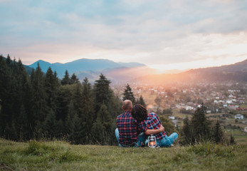 happy loving couple sitting hugging on a grass on a top of mountain at magical sunset. Man and woman traveling together. Traveler couple enjoying nature, embracing, watching landscape under a blanket.