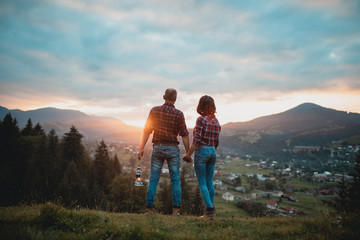 couple standing arm in arm on a mountain rock. against a background of a sunset in the mountains. A romantic date. Back view. Banner