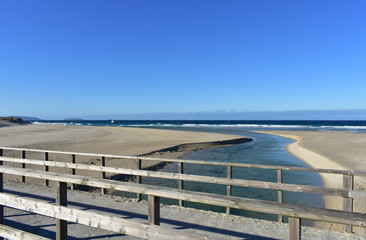Fototapeta na wymiar Beach with river, wooden handrail and seagulls. Golden sand and sea with waves and white foam. Blue sky, sunny day. Galicia, Coruña Province, Spain.