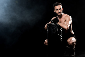 Fototapeta na wymiar shortless boxer sitting and holding punching bag on black with smoke