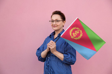 Eritrea flag. Woman holding Eritrean flag. Nice portrait of middle aged lady 40 50 years old holding a large flag over pink wall background on the street outdoor.