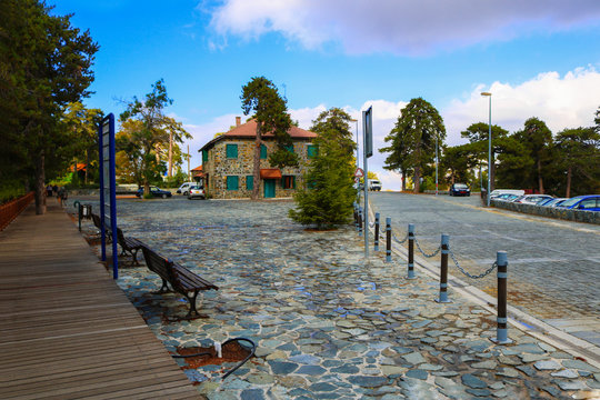 Tourist place in Trodos Mountains, Cyprus. View on pedestrian road made of stones, car parking, and two storied house, pine trees and bright sky. For wallpaper, postcart and design. Horizontal