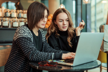 Two girls, friends Drink Hot Coffee While Work In Cafe On Laptop. Portrait Of Stylish Smiling Girls In Winter Clothes Drinking Hot Coffee And Work At Laptop. Female Winter Style. - Image