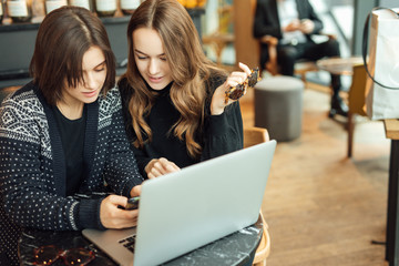 Two girls, friends Drink Hot Coffee While Work In Cafe On Laptop. Portrait Of Stylish Smiling Girls In Winter Clothes Drinking Hot Coffee And Work At Laptop. Female Winter Style. - Image