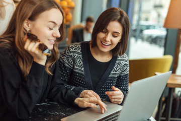 Two girls, friends Drink Hot Coffee While Work In Cafe On Laptop. Portrait Of Stylish Smiling Girls In Winter Clothes Drinking Hot Coffee And Work At Laptop. Female Winter Style. - Image