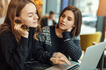 Two girls, friends Drink Hot Coffee While Work In Cafe On Laptop. Portrait Of Stylish Smiling Girls In Winter Clothes Drinking Hot Coffee And Work At Laptop. Female Winter Style. - Image