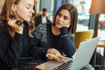 Two girls, friends Drink Hot Coffee While Work In Cafe On Laptop. Portrait Of Stylish Smiling Girls In Winter Clothes Drinking Hot Coffee And Work At Laptop. Female Winter Style. - Image