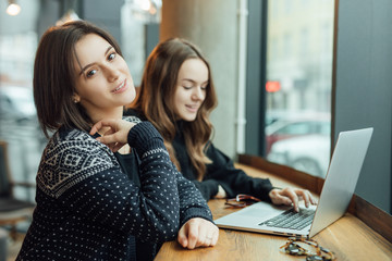 Two girls, friends Drink Hot Coffee While Work In Cafe On Laptop. Portrait Of Stylish Smiling Girls In Winter Clothes Drinking Hot Coffee And Work At Laptop. Female Winter Style. - Image