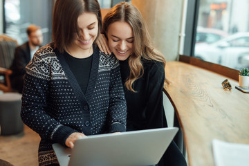 Two girls, friends Drink Hot Coffee While Work In Cafe On Laptop. Portrait Of Stylish Smiling Girls In Winter Clothes Drinking Hot Coffee And Work At Laptop. Female Winter Style. - Image