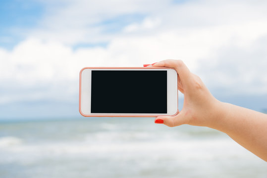 Woman hand showing a blank smart phone horizontal screen display on the beach with the sea in the background