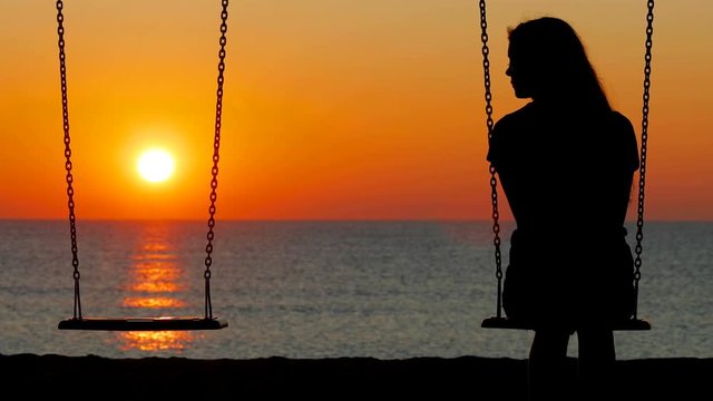 Back view silhouette of a sad girl swinging looking at side t an empty seat on the beach at sunset