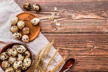 quail eggs in a wooden bowls and plates with broken eggshell on a rustic table linen with ears of wheat and wooden spoon on rustic background