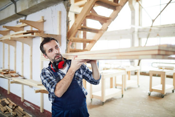 Serious handsome bearded builder in overall wearing ear protectors on neck walking over construction site and carrying wooden planks on shoulder