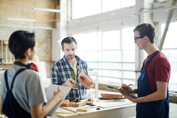 Serious skilled middle-aged carpenter standing at desk and showing wooden pieces to students while holding class in workshop
