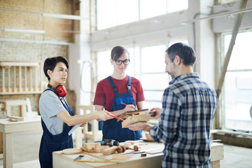 Curious young carpenters in overalls standing at desk with wooden details and making notes in workbooks while asking questions to foreman during class in carpentry shop.