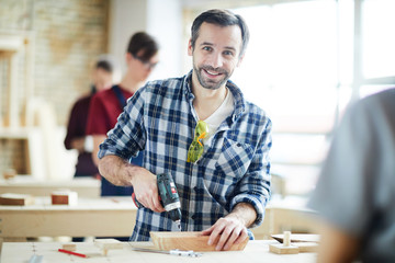 Happy handsome bearded man in checkered shirt standing at table and drilling wooden piece with power tool while looking at camera
