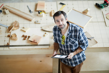 Content handsome middle-aged man with beard frowning forehead and holding tablet while leaning on table with wooden details and looking at camera in carpentry