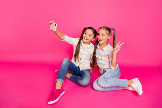Close up photo two people little age she her girls hold hands arms telephones make take selfies v-sign sit floor  wear casual jeans denim checkered plaid shirts isolated rose vibrant background