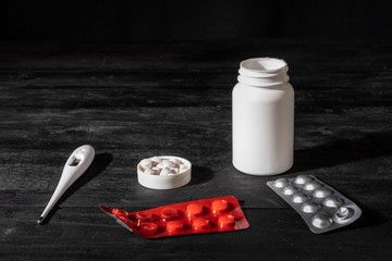 Medical pills and thermometer on black wood background. Self treatment concept: minimalistic low key image of prescription drugs.