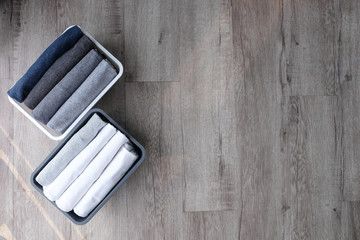 Stack of folded t shirt black gray white color in a plastic basket on wooden floor background in natural light with copy space. Room cleaning and tidying up concept. 