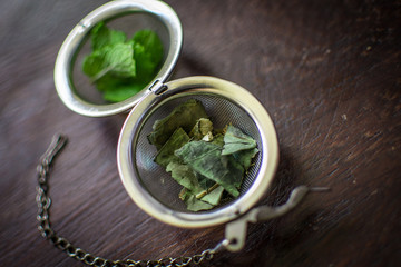 Metal tea infuser with dried leaves of green tea and fresh mint on a dark brown wooden background