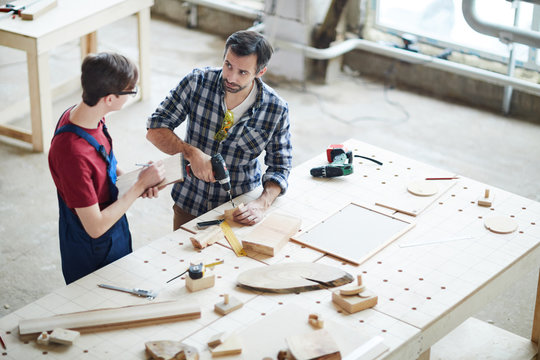 Serious confident handsome middle-aged man with beard standing at desk and drilling wooden detail while showing intern with sketchpad how to make hole in wooden piece.