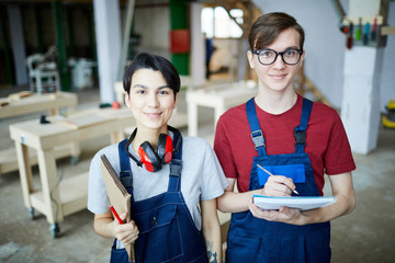 Content young carpentry students in overalls standing in modern workshop and looking at camera while visiting woodworking course
