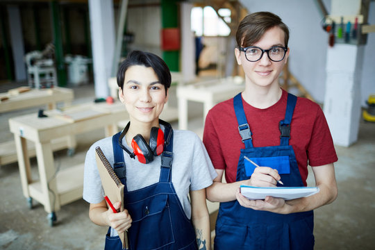 Content young carpentry students in overalls standing in modern workshop and looking at camera while visiting woodworking course