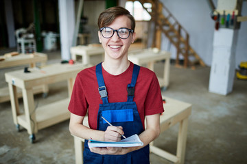 Jolly excited young guy in glasses and overall making notes in workbook and looking at camera while...