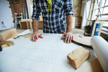 Close-up of unrecognizable male engineer standing at table with blueprint and analyzing it while working on house project in own workshop