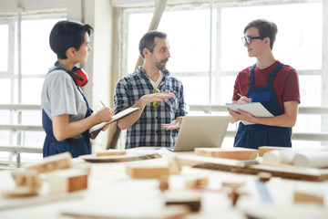 Confident handsome chief carpenter in checkered shirt gesturing hands while giving class for interns in workshop, young students making notes in sketchpads