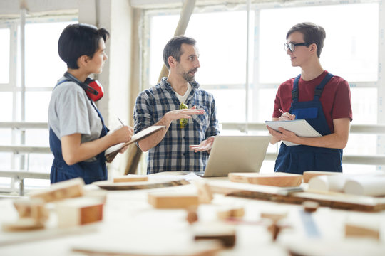 Confident handsome chief carpenter in checkered shirt gesturing hands while giving class for interns in workshop, young students making notes in sketchpads