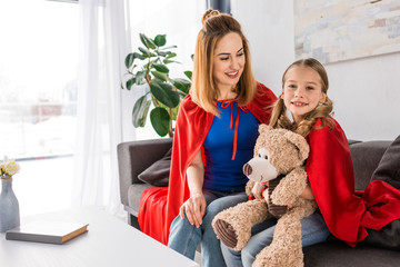 Beautiful mother and kid in red cloaks holding teddy bear and sitting on sofa at home
