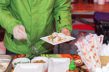 Chef makes portion of the food for his customer, paper coaster with dumplings and vegetable decoration in one hand, spoon in the other hand. Asian street food market, fast food. Motion blur.