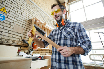 Cheerful handsome bearded carpenter in protective goggles standing in workshop and using triangle...
