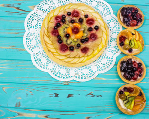 one large fruit tartlet and four small ones for a light snack on a blue background with apples, pears, kiwi, grapes, raspberries and strawberries. Healthy and very satisfying food.