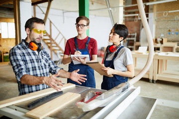Serious confident skilled carpenter in safety goggles gesturing hands while pointing at circular...