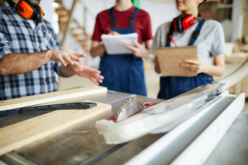 Close-up of unrecognizable male carpenter gesturing hands while showing circular saw table to students during carpentry class at factory