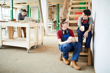 Positive young workers in overalls sitting on wooden stairs in workshop and talking while drinking...