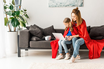 mother and daughter in red cloaks sitting on sofa and holding laptop