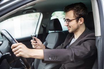 Young guy is sitting at his car. He is looking at the keys in his right hand. His left hand is on the steering wheel.