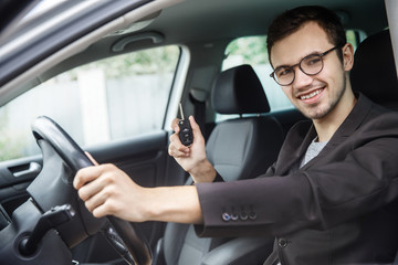 Young man is sitting at his car while looking at the camera. He is holding the keys in his right hand. His left hand is on the steering wheel.