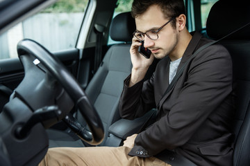 Young guy clips on his belt while talking on the phone. He is sitting at his car. Road safety concept.