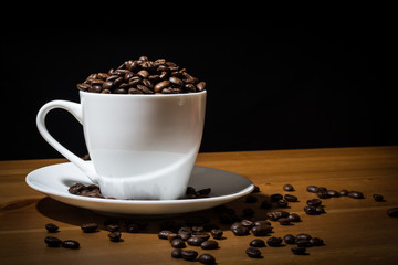 White cup filled with coffee beans  and coffee beans scattered on a wooden surface and on the white saucer. 